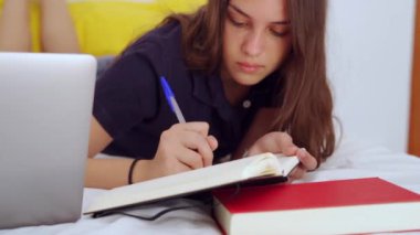 Genç kız öğrenci evde uzaktan eğitim sırasında bilgisayarla okurken yatakta uzanıyor ve deftere yazı yazıyor.