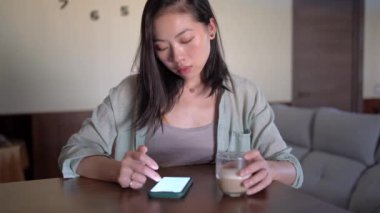 Evdeki masada cep telefonuyla internette bir bardak kahve sörfü yapan genç etnik bir kadın.