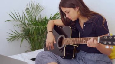 Evde boş zamanlarında akustik gitar çalmayı öğrenirken dizüstü bilgisayarda video dersi izleyen kız öğrenci.