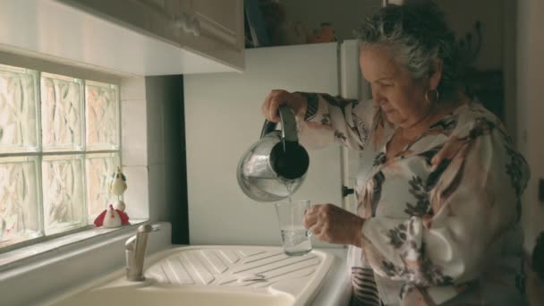 聚精会神的老年妇女一边在厨房里用茶袋做饮料 一边用透明电水壶把热水倒入玻璃杯的侧视图 — 图库视频影像
