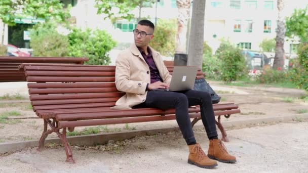 自信を持って若いヒスパニックの男性は スタイリッシュなカジュアルな服装とメガネは ベンチに座って市街地のラップトップで作業しながら電話に応答します — ストック動画