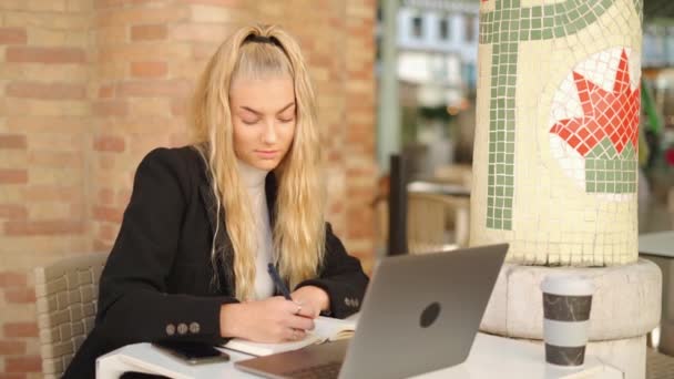 ラップトップとコーヒーでテーブルに座ってカフェで働いている間 プランナーでメモを取る集中した女性フリーランサー — ストック動画