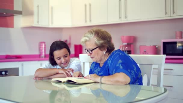 全神贯注的祖母穿着休闲装和眼镜坐在桌旁 和快乐的孙女一起在厨房里看书 — 图库视频影像