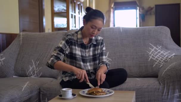 リビングルームで快適なソファーに座っている間 テーブルの上のコーヒーのカップ近くのプレートに置かれた自家製パンケーキを食べる若いアジアの女性 — ストック動画