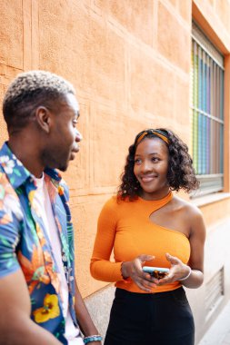 Genç siyah çift, güneşli bir günde şehirde yürürken günlük ve renkli kıyafetler giyip birbirleriyle konuşuyorlar.