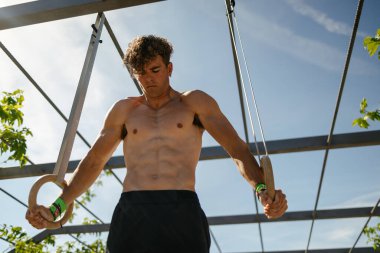 Sarışın, yakışıklı genç adam güneşli bir günde dışarıda yüzüklerle egzersiz yapıyor.