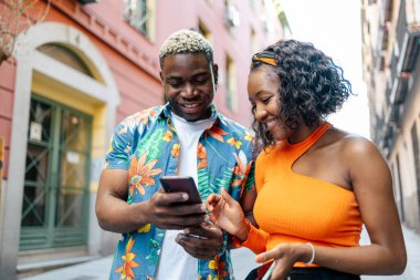 Renkli ve günlük giysiler içinde genç siyah bir çift akıllı telefonlarını sokakta kullanırken şehirde dolaşıyorlar. Adam, akıllı telefondaki kadına gülümserken bir şey gösteriyor.