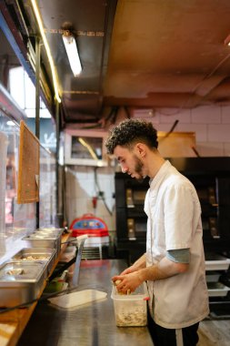 Beyaz önlüklü genç Arap pizzacı pizzacının mutfağındaki bir kutuya mantar koyuyor.