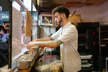 Beyaz önlüklü genç Arap pizzacı pizzacının mutfağındaki bir kutuya mantar koyuyor.
