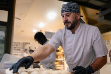 Beyaz elbiseli ve siyah şapkalı bir fırıncı hamur topları yapıp bir fırının tepsisine koyuyor.