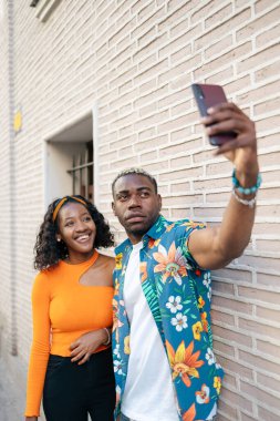 Genç siyah çift gündelik ve renkli kıyafetler giyip şehirde yürürken selfie çekiyorlar ve gülümsüyorlar.