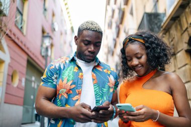 Renkli ve günlük giysiler içinde genç siyah bir çift akıllı telefonlarını sokakta kullanırken şehirde dolaşıyorlar.