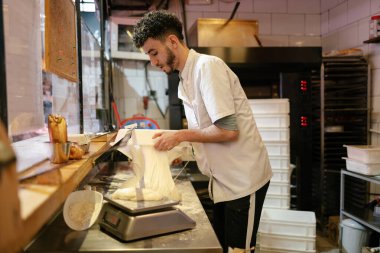 Beyaz önlüklü genç Arap pizzacı, pizzacının mutfağındaki masada çalışmak için plastik bir kabın içindeki hamuru çıkarıyor.