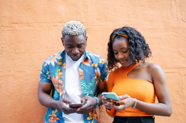 Her ikisi de akıllı telefonlarını kullanırken renkli ve sıradan elbiseli genç bir çift duvara yaslanıyorlar.