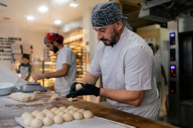 Beyaz elbiseli ve siyah şapkalı yetişkin bir fırıncı, Arap bir meslektaşla mangır topları yapıp fırının arka planında hamur kesiyor.