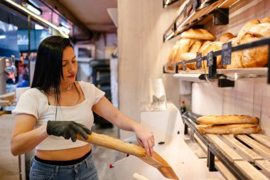 Siyah saçlı ve beyaz tişörtlü genç Latin bir kadın fırında müşteri için eldivenli bir somun ekmeği çantaya koyuyor.