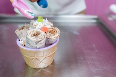 A worker is decorating a rolled ice cream tub with chips and teddy bear toppings with a spoon. Concept of new small business clipart