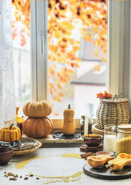 Autumn kitchen still life with various pumpkins, kitchen utensils and ingredients at concrete table with window background with fall foliage