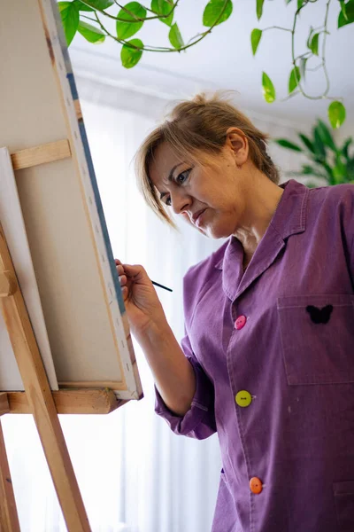 Adult woman with blonde hair dressed in a purple robe paints with brush on canvas next to the window in her home