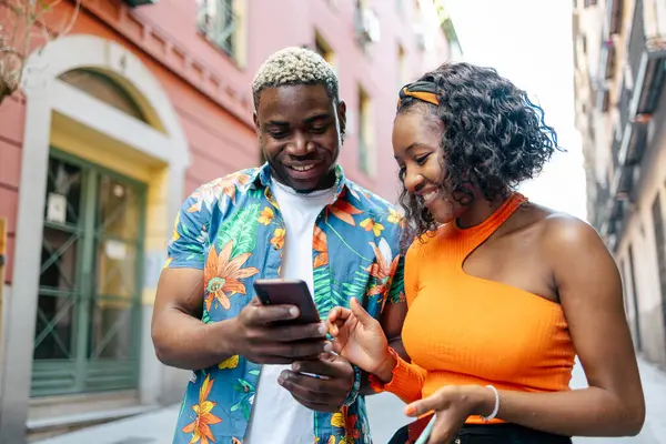 Young black couple in colorful and casual clothes walking around the city while using their smartphone in the street. The man shows something to the woman on the smartphone while smiling