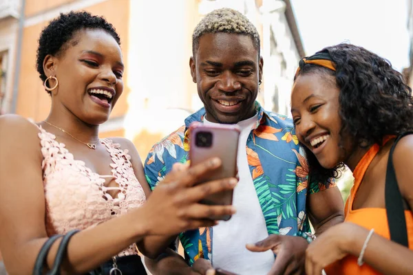 Three young black friends meeting on the street and walking around the city while using their smartphones. One of the women is showing something to her friends