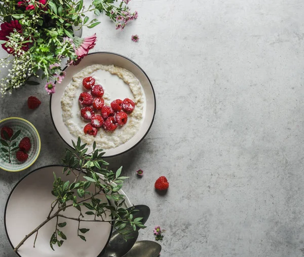 Romantic breakfast with porridge and heart of raspberries on grey kitchen table with flowers and bowls. Vegan breakfast idea with nutritious ingredients. Top view with copy space.