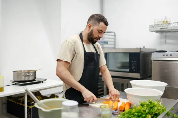 Young bearded chef dressed in a kitchen apron cutting carrots in the table of a well-equipped kitchen