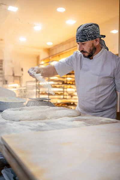 Caucasian adult baker in white apron and black cap pouring flour on the dough to work it on a table in a bakery