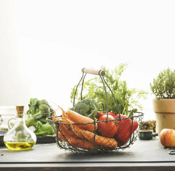 Metal grocery basket with organic vegetables: carrots, broccoli, bell pepper and tomatoes on table with other healthy ingredients at window background with sunlight. Healthy lifestyle. Front view.