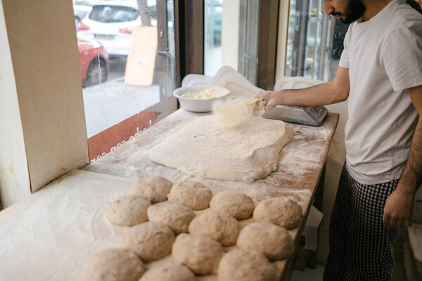 Arabic young baker in white t-shirt pouring flour on the dough with loaves of dough next to it on a table in a bakery