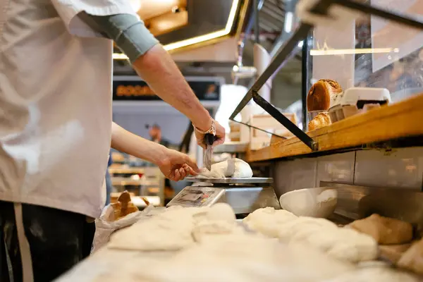 Detail shot of a man in white apron weighing dough on a scale while cutting it with the scapula in a bakery