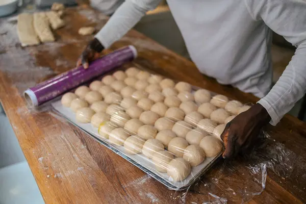 Portrait of a multitude of small balls of dough on a tray on a table while a baker covers them with plastic in a bakery