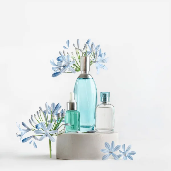 Mavi cam spreyli güzellik ürünleri ve podyumda beyaz arka planda çiçek ve taç yapraklarıyla duran pipo şişeleri. Serum, parfüm kokusu ve tonikli kozmetik seti. Ön görünüm