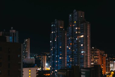 Şehrin yerleşim bölgesinde gece gökyüzüne karşı parlayan pencereleri olan uzun apartmanlar.