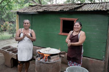 Two adult Hispanic women are smiling while shaping corn tortillas by hand and using an outdoor stove in Guatemala clipart