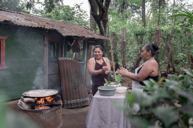 İki yetişkin İspanyol kadın gülümsüyor ve mısır tortillalarını elle şekillendiriyor ve Guatemala 'da açık hava sobası kullanıyorlar.