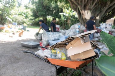 Ön cephede plastik ve kartonla dolu dolu dolu bir el arabası dururken, arka planda işçiler geri dönüşüm faaliyetlerinde çalışırken,