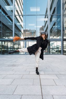 Tüm vücudu açık giyinen, buketli, neşeli genç bir kadın. Şehrin sokaklarında camdan duvarları olan modern bir binaya karşı kaldırımda dans ederken gülümseyip bacağını kaldırıyor.