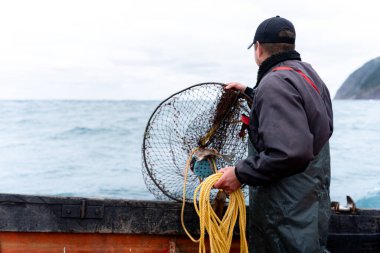 Rear view of a caucasian young fisherman picking up a container with nets for lobster fishing from the water clipart