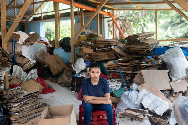 портрет серьезной молодой испаноязычной женщины среди склада, заполненного картонной коробкой для переработки отходов, проецирующей решимость и серьезность в своей приверженности к экологической ответственности