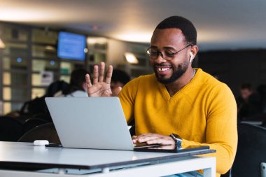 Süveter ve gözlük takan neşeli Afrikalı Amerikalı sakallı erkek girişimci ofiste çalışırken kulaklıkla netbook üzerinden video görüşmesi yapıyor.