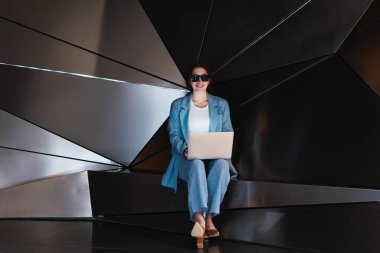 Güneş gözlüklü çekici bir kadın netbook 'ta internette sörf yaparken modern stüdyoda yaratıcı 3 boyutlu duvarlarla oturuyor.