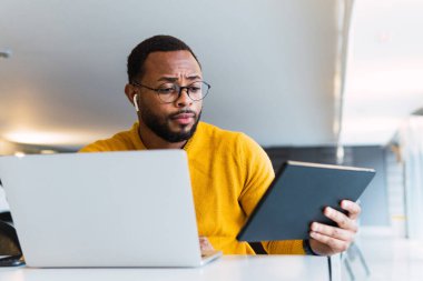 Düşünceli siyah erkek, dizüstü bilgisayarla masada oturuyor ve birlikte çalışma alanındaki tabletten kulaklıkla müzik dinliyor.