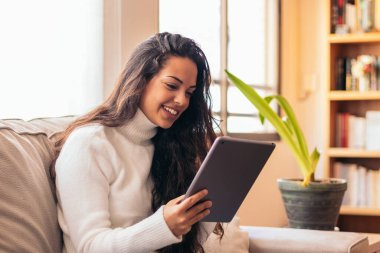 Evindeki kanepede oturan genç bir kadın, rahat kıyafetler giymiş bir tablet kullanıyor.