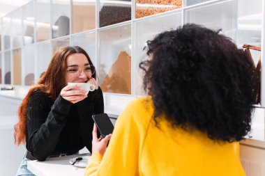 Modern kafede isimsiz etnik bir bayan arkadaşıyla sohbet ederken gülümseyen ve kahve içen gündelik kıyafetleri ve gözlükleriyle uzun kızıl saçlı pozitif bir genç bayan.
