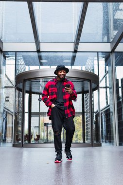 Şapkalı ve kareli tişörtlü Afro-Amerikan adam cam duvarlı ve cep telefonlu bir binanın lobisinde yürüyor.