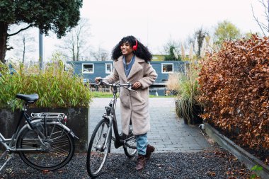 Kıvırcık siyah saçlı, sırıtan genç bir Afro-Amerikan kadın. Sıcak bir palto giyiyor, bisikletle yürüyor ve sonbahar günü sokakta kulaklıkla müzik dinliyor.