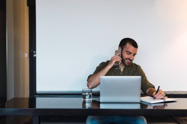 Çalışma sürecinde masa başında oturmuş modern netbook ve dizüstü bilgisayarla telefon görüşmesi yapan pozitif erkek işçi.