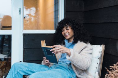 Yandan bakan genç Afro-Amerikan kadın sıcak kıyafetli ilginç bir kitap okuyor ve gün ışığında terasta rahat bir sandalyede dinleniyor.