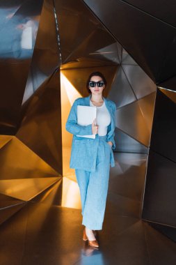 Güneş gözlüklü, modern netbook 'lu modern 3D geometrik duvarlı stüdyoda yürüyen şık bir kadın vücudu.